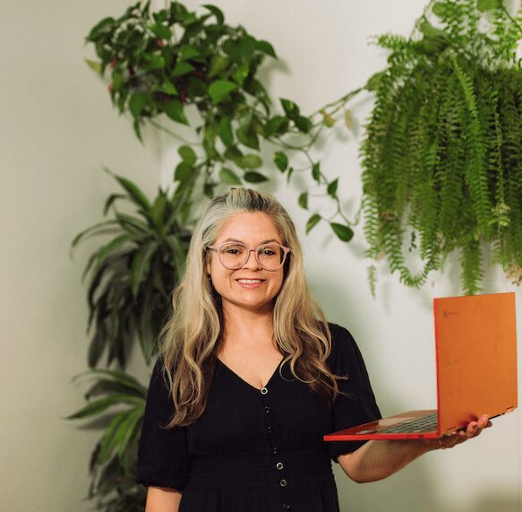 Smiling white woman holding a laptop in front of several plants