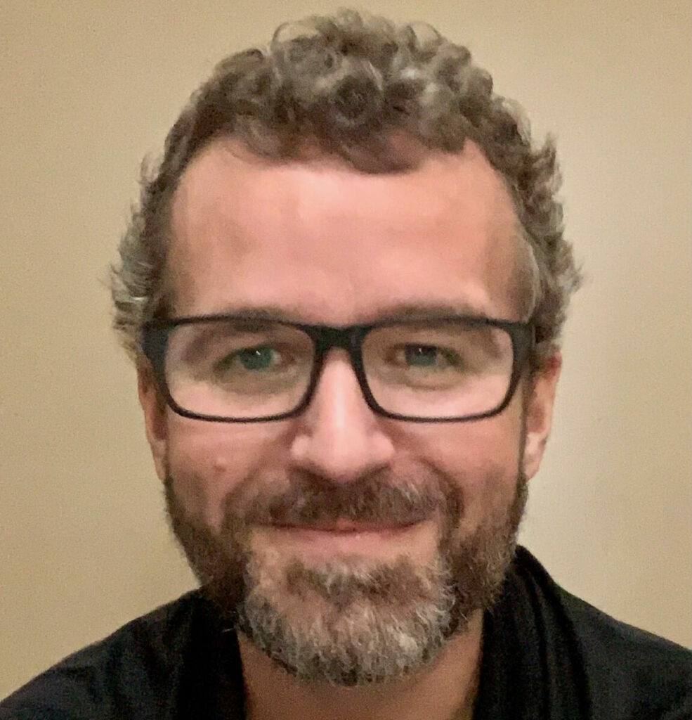 A white man with glasses and a black shirt smiling